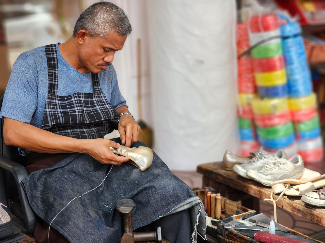 Artisan hand-stitching a gold-toned shoe beside cobbler tools and an anvil