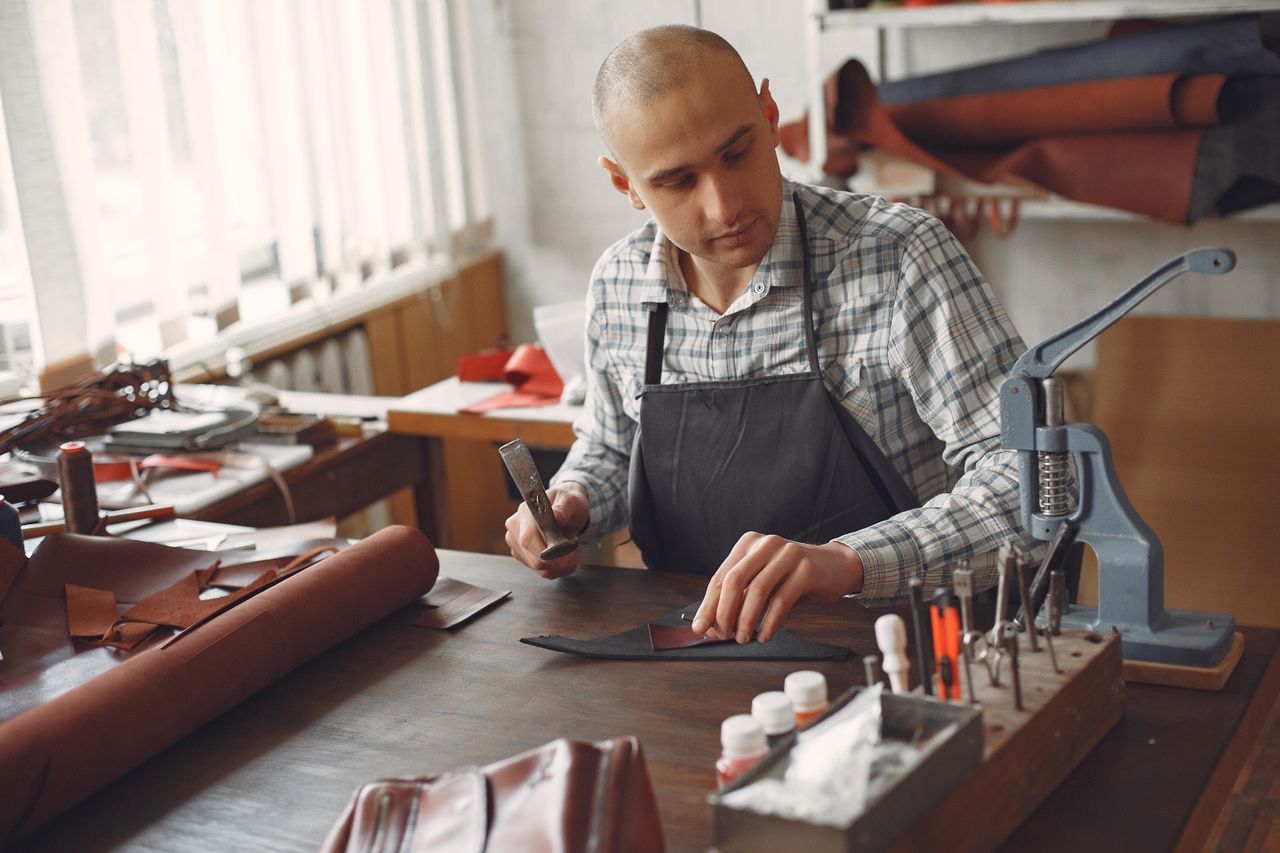 Leatherworker using hand tools and a press at a sunlit workshop table with leather rolls